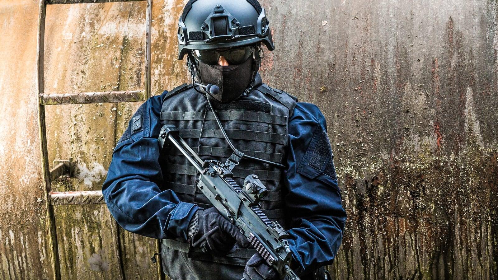 Operator in blue uniform and plate carrier holding an MP7 in front of a weathered steel tank