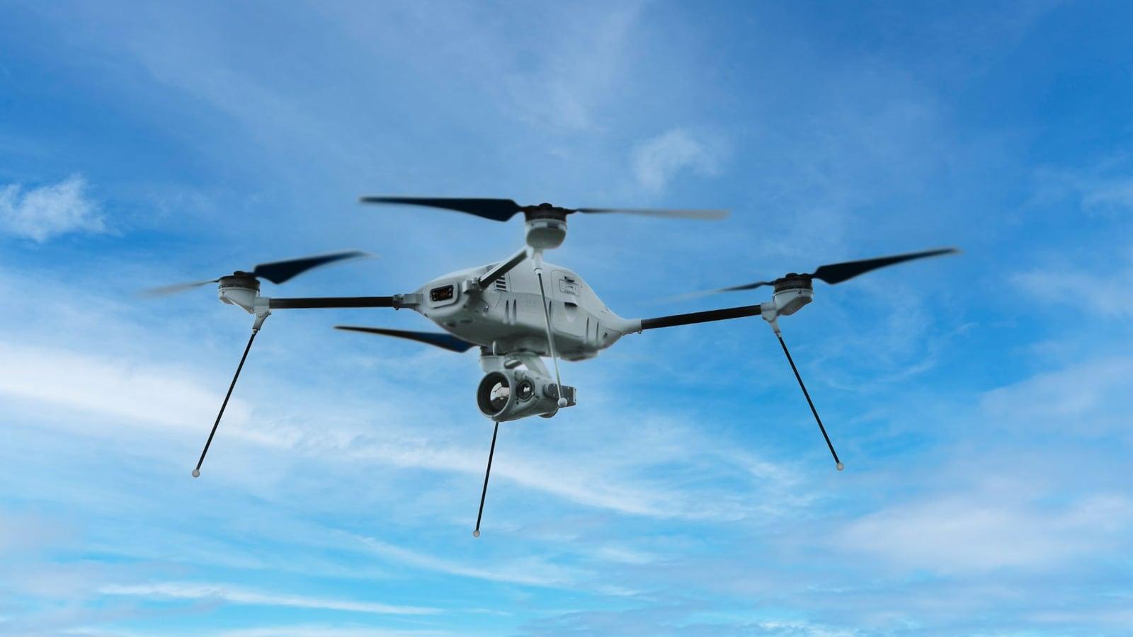 A grey four-rotor drone with a gimballed camera payload hovering against a blue sky