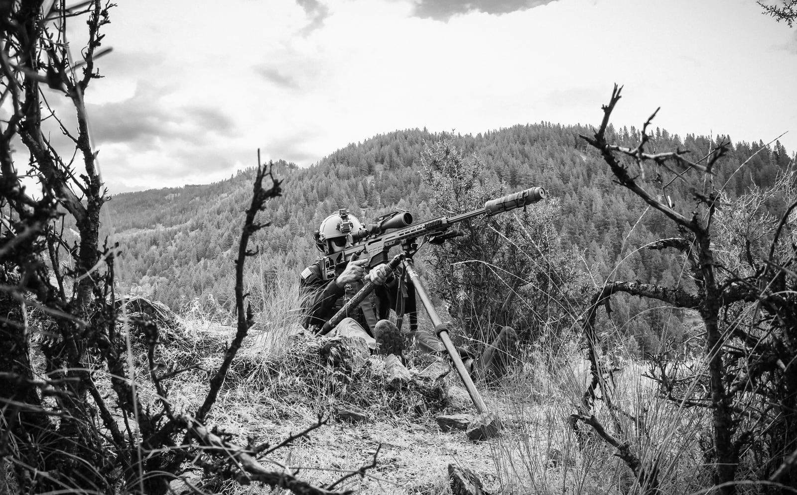 Sniper firing the MK 22 from a tripod on a brushy hillside, black and white