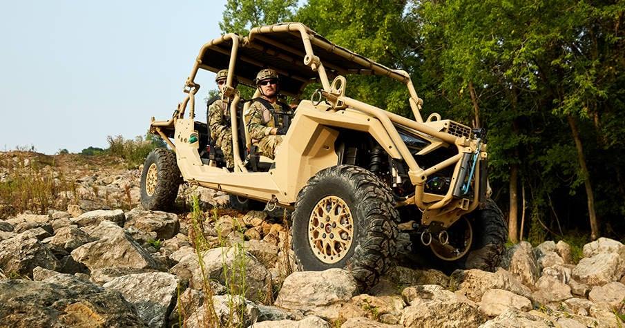 Tan MRZR Alpha with crew climbing over rocks at the edge of a forest