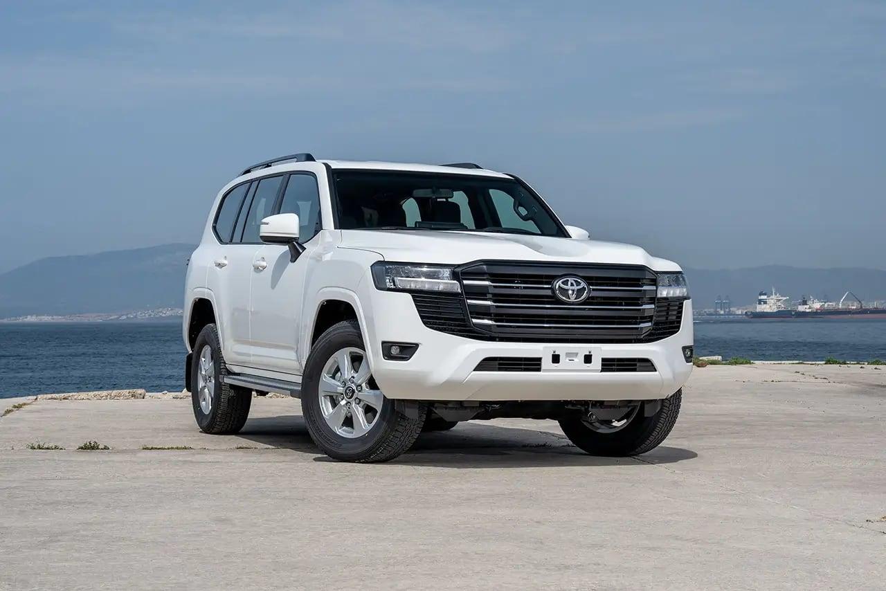 A white Land Cruiser 300 SUV parked on a concrete quay with the sea and hills behind