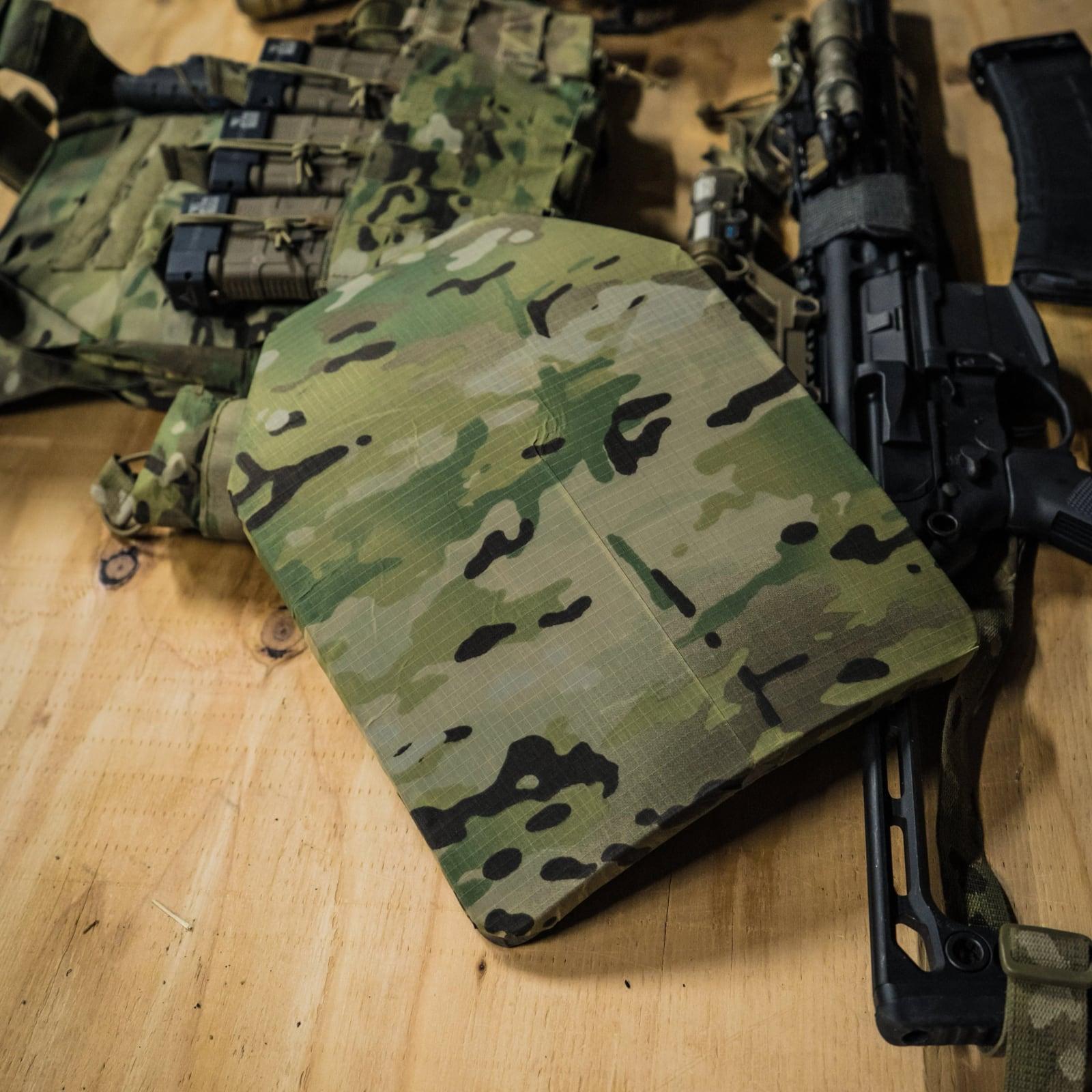 Multicam-wrapped plate on a workbench beside a plate carrier and rifle