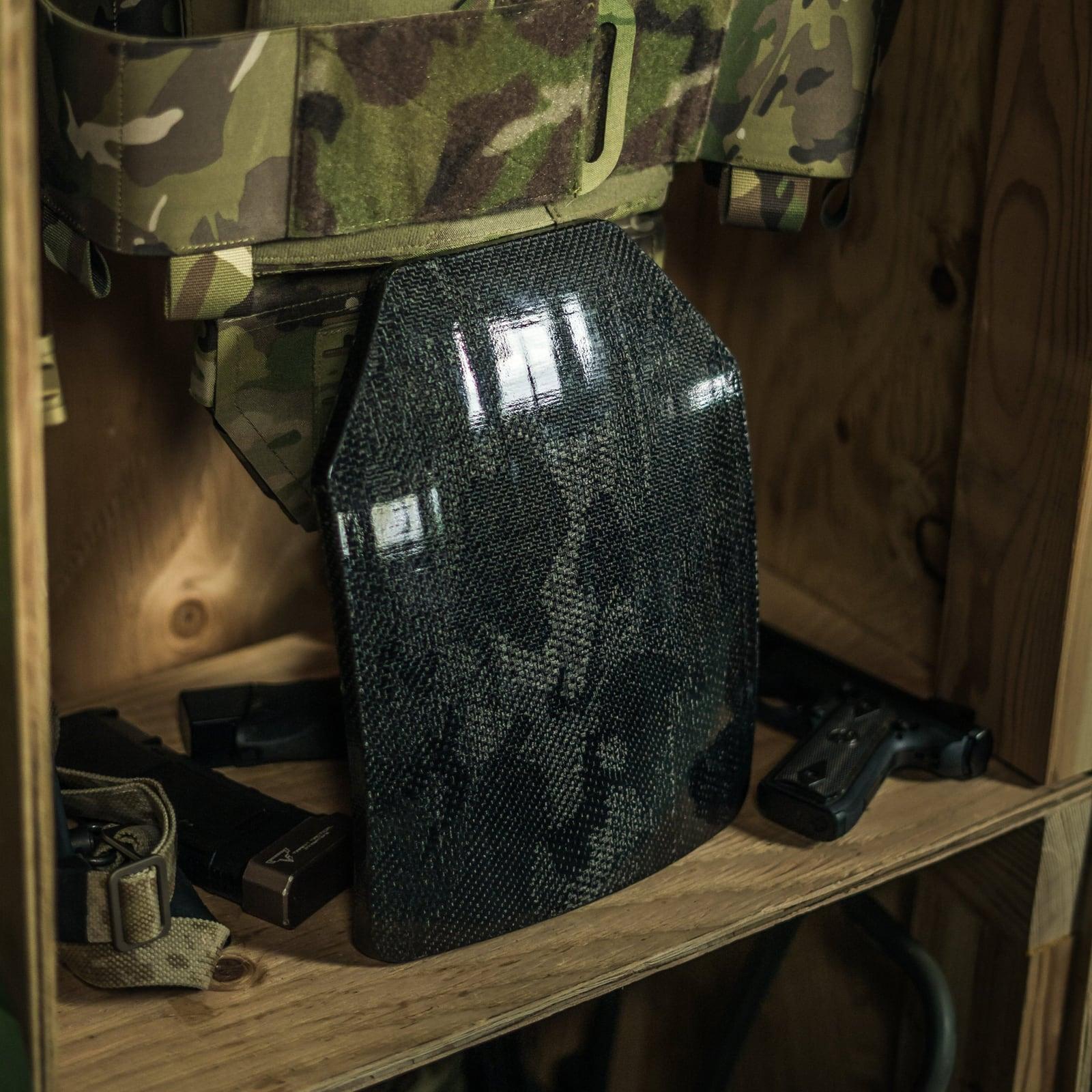 Black US Armor Forge MK-V plate standing on a wooden shelf in front of a camouflage plate carrier